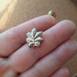 May include: A silver-toned metal lapel pin shaped like a stylized oak leaf with five distinct lobes. The surface of the leaf features embossed vein details and a central stem, with small dark spots visible on the tips of the leaf lobes.