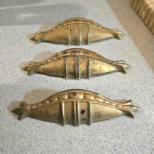 May include: Three antique brass drawer pulls with a decorative, symmetrical design. Each pull has a curved shape with vertical and horizontal lines, and a slightly aged appearance. The pulls are arranged in a vertical row.