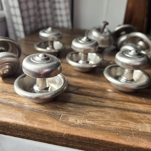 May include: A collection of antique silver-toned drawer pulls, each with a circular top and a small, bowl-shaped base. The pulls are arranged on a wooden surface, showcasing their vintage design and potential for furniture restoration or decorative projects.