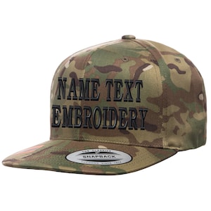 May include: A camouflage green and brown baseball cap with black embroidered text that reads "NAME TEXT EMBROIDERY". The cap has a black snap closure and a label that reads "THE CLASSICS SNAPBACK YUPOONG SINCE 1974".