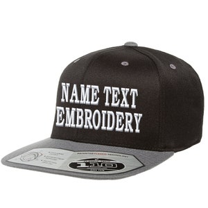 以下が含まれることがあります： 「NAME TEXT EMBROIDERY」の文字が白で刺繍された、黒とグレーのフラットブリムベースボールキャップ。キャップはグレーのつばと上部にグレーのボタンが付いています。Flexfitテクノロジーのラベルがつばに表示されています。