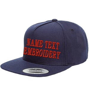 Puede incluir: Una gorra de béisbol azul marino con texto bordado en rojo que dice "NAME TEXT EMBROIDERY". La gorra tiene cierre a presión y un aspecto clásico.