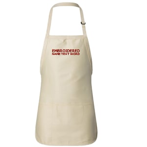 Puede incluir: Un delantal de algodón beige con una correa de cuello ajustable y un bolsillo grande. El delantal tiene un texto bordado en rojo que dice "EMBROIDERED NAME TEXT WORD".