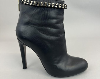 May include: Black leather ankle boots with a gold chain detail around the top of the boot. The boots have a stiletto heel.