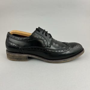 Może przedstawiać: Czarny skórzany but typu brogue z dekoracyjnymi perforacjami i niskim obcasem. But ma klasyczny design ze skrzydłowymi noskami i sznurowane zapięcie. Podeszwa ma postarzany wygląd.