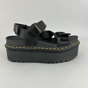 May include: Black platform sandals with adjustable straps. The sandals feature a thick, ridged sole with yellow stitching around the edge. The upper straps are made of a textured material, and the sandals have a modern, chunky design.