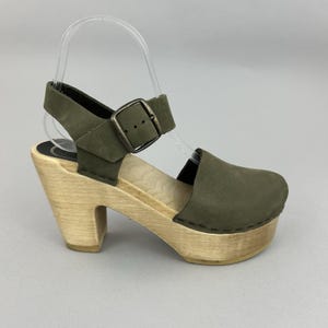 May include: Olive green platform clog sandal with a wooden sole and heel. The sandal features a buckle closure and a strap around the ankle. The heel measures approximately 3 inches.