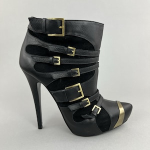 May include: Black leather ankle boot with multiple straps and gold-coloured buckles. The boot features a pointed toe and a high stiletto heel. The design includes cut-out details and a metallic accent on the toe.