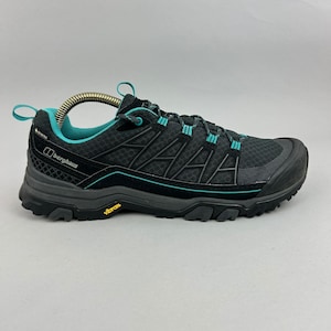 May include: A black Berghaus hiking shoe with teal accents, featuring a mesh upper, a Gore-Tex label, a dark grey midsole, and a yellow Vibram logo on the rugged outsole. The shoe includes a pull tab at the heel and a lace-up closure system.