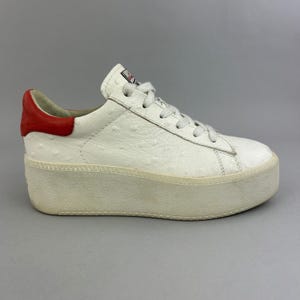 Può includere: Una sneaker bassa bianca con una texture effetto pelle di struzzo, dotata di una spessa suola platform, lacci bianchi e un inserto a contrasto in pelle rossa sul tallone. La linguetta presenta una piccola etichetta rettangolare con testo rosso e nero.