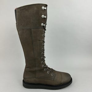 May include: A tall, brown leather boot with a lace-up design and a flat sole. The boot features silver-tone metal accents and a high shaft that extends to the knee. The boot is approximately 38 cm tall.