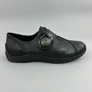 May include: A black slip-on shoe featuring a smooth leather front, a textured heel panel, and a decorative silver-toned spiral button accent on a wide strap across the instep. The shoe has a low profile, a rounded toe, and a black rubber sole.