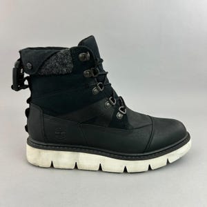 May include: A black ankle boot featuring a smooth leather toe cap, a textile upper, and a fold-over collar with a grey wool-like lining. The boot includes black laces with metal eyelets, a side buckle detail, and a thick, white, lugged rubber sole.