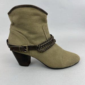 Può includere: Stivali da caviglia in tela beige con cinturino in pelle marrone e dettaglio fibbia. Gli stivali hanno un tacco spesso e un accento a catena intrecciata.