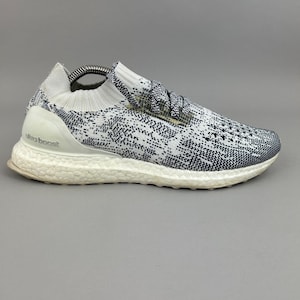 May include: A white and black knit sneaker featuring a textured pattern on the upper, a white ribbed collar, and a white foam midsole with the text ultra boost embossed on the heel counter. The shoe includes white laces and a white outsole.