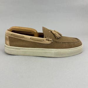 May include: A tan suede loafer with a cream-colored sole. The shoe features a tassel detail and a leather trim. The shoe is designed with a slip-on style and is likely for casual wear.