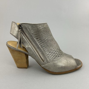 Könnte beinhalten: Silberne Peep-Toe-Sandale mit gewebtem Design am Obermaterial und seitlichem Reißverschluss. Der Schuh hat einen Holzblockabsatz und ein kleines Riemendetail am Knöchel. Die Sohle ist hellbraun.