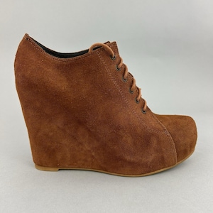 May include: Brown suede wedge ankle boots with a lace-up design. The boots have a rounded toe and a high wedge heel. The suede material has a soft, textured appearance. The boots are a warm, earthy tone.