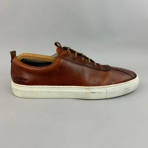May include: A low-top sneaker crafted from smooth brown leather with a rounded toe, tonal brown laces, and a thick white rubber sole. A wooden shoe tree is inserted inside the shoe, and a small embossed logo is visible on the heel area.