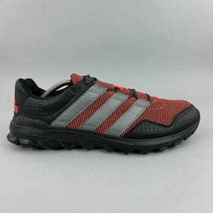Può includere: Scarpa sportiva nera e rossa con strisce grigie. La scarpa presenta una suola e dettagli neri, con il logo Adidas sul lato. I lacci sono neri e rossi.