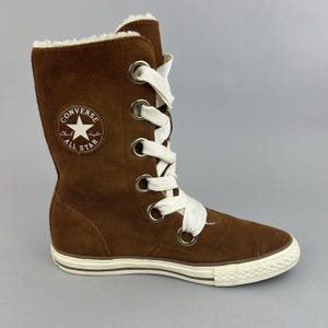 Op de afbeelding: Bruine suède Converse All Star hoge laars met witte veters en een crèmekleurige zool. De laars heeft een imitatiebontvoering en de Converse All Star-logopatch. De laars is halverwege de kuit.