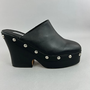 Op de afbeelding: Een zwarte instapmuiltje met een dikke plateauzool en een hoge blokhak. De schoen is voorzien van een rij zilverkleurige metalen studs langs de basis van het bovenwerk. Een zwart label met witte tekst is zichtbaar op het voetbed aan de binnenkant.