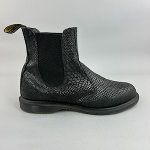 May include: Black ankle boot with a textured, snake-like pattern. The boot features a rounded toe, a black sole, and elastic side panels for easy wear. A yellow tag with black text is attached to the back of the boot.