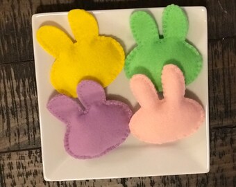 May include: Four felt bunny shapes in different colors: yellow, green, purple, and pink. The bunnies are on a white plate.