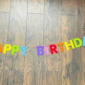 May include: A colorful felt banner with the words "Happy Birthday" strung on a white string. The banner is hanging on a wooden floor.