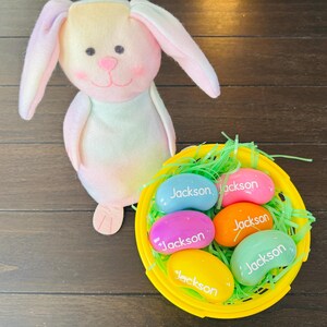May include: A yellow basket filled with colorful jelly beans, each with the name "Jackson" written on them. The basket is sitting on a wooden surface next to a pastel-colored stuffed bunny.