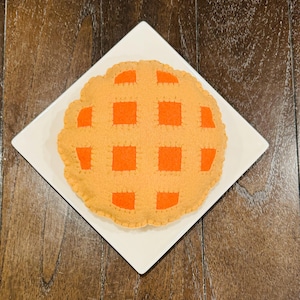 May include: A felt pie with a brown crust and an orange filling. The pie is on a white plate.