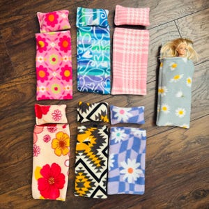 May include: A collection of doll-sized sleeping bags and pillows in various patterns and colors. Designs include floral, plaid, and geometric prints. One sleeping bag contains a doll. The items are made of soft fabric.