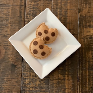 May include: Two felt chocolate chip cookies on a white plate. The cookies are brown with dark brown chocolate chips.