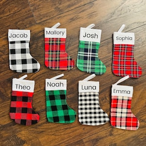 May include: Eight personalized Christmas stockings with different plaid patterns. The stockings are red and green plaid, black and white plaid, and red and black plaid. Each stocking has a white cuff with a name written in black.