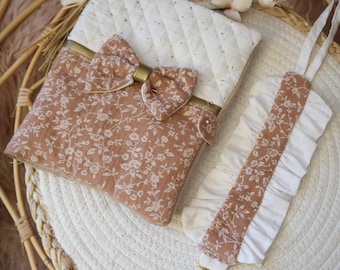 May include: A baby accessory set featuring a quilted cream-colored changing pad cover and a matching pacifier clip. The set includes a floral print in shades of brown and white, with a decorative bow and gold accents.