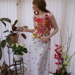 May include: A white maxi dress with a pink and brown floral print. The dress has a tie at the neckline and a fitted bodice. The model is standing in a sunroom with plants.