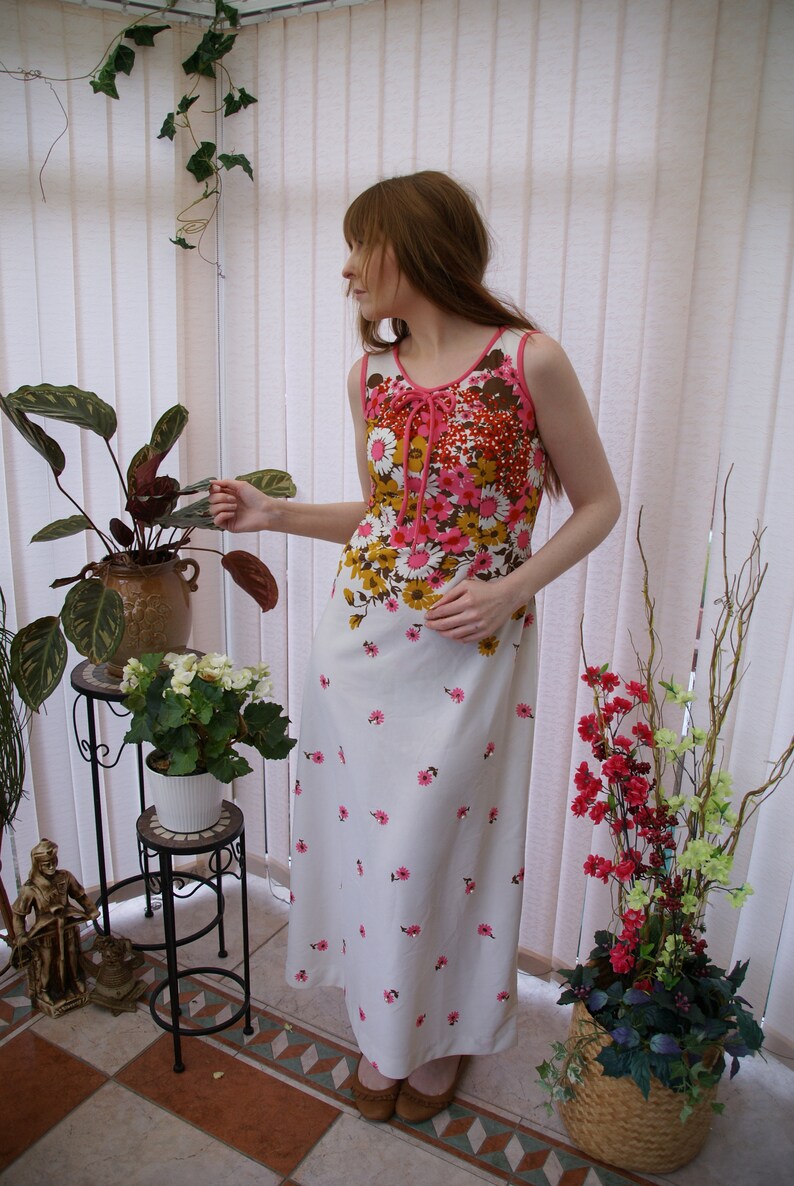 May include: A white maxi dress with a pink and brown floral print. The dress has a tie at the neckline and a fitted bodice. The model is standing in a sunroom with plants.