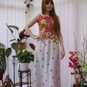 May include: A white maxi dress with a floral print in shades of pink, brown, and yellow. The dress has a sleeveless bodice and a fitted waist. The model is wearing the dress in a greenhouse setting.
