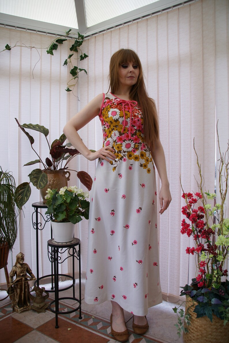 May include: A white maxi dress with a floral print in shades of pink, brown, and yellow. The dress has a sleeveless bodice and a fitted waist. The model is wearing the dress in a greenhouse setting.