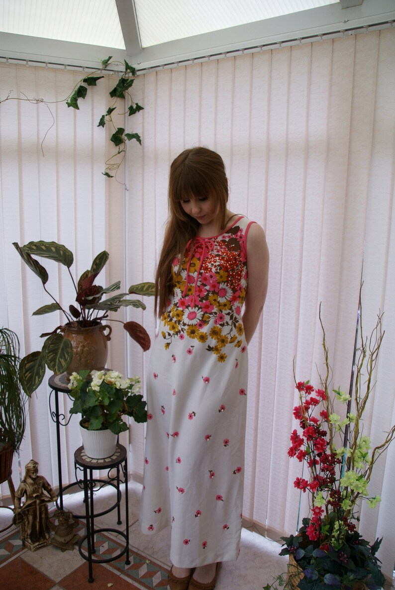 May include: A woman wearing a white dress with pink and yellow floral print. The dress has a scoop neck and sleeveless design. She is standing in a sunroom with plants and white blinds.