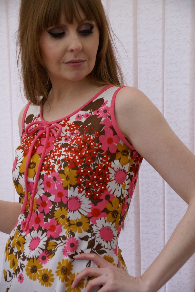 May include: A sleeveless dress with a pink and white floral print. The dress has a pink bow at the neckline and is adorned with small white beads.