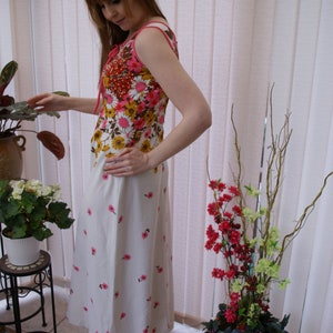 May include: A woman wearing a white maxi dress with pink and brown floral print. The dress has a sleeveless bodice and a fitted waist. She is standing in front of a window with white blinds.