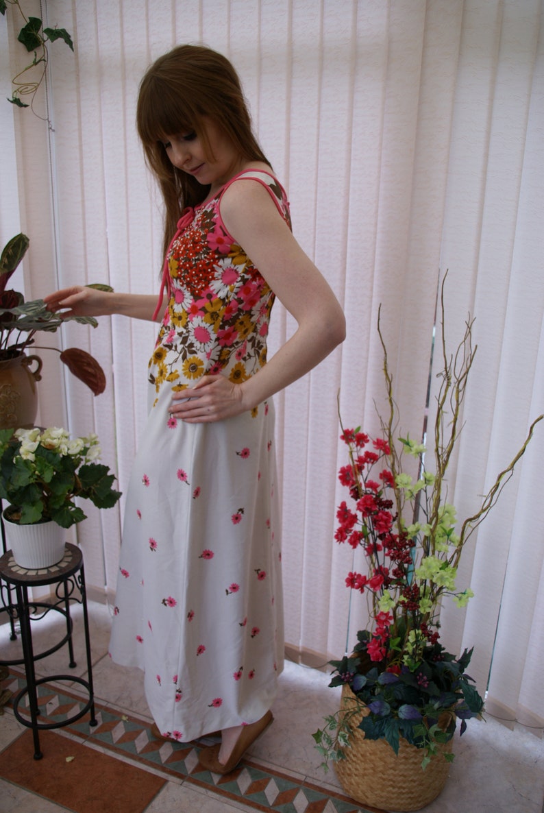 May include: A woman wearing a white maxi dress with pink and brown floral print. The dress has a sleeveless bodice and a fitted waist. She is standing in front of a window with white blinds.