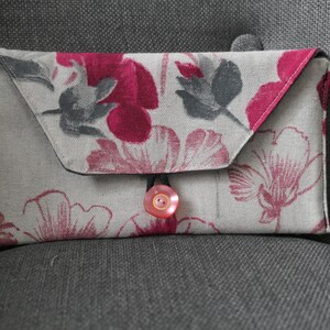 Puede incluir: Un pequeño bolso de mano con estampado floral y cierre de botón rosa. La tela es de color beige claro con diseños florales rosas y grises.