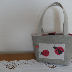 Può includere: Una piccola borsa tote in lino grigia con una tasca che presenta un design di coccinella rossa e nera. La borsa ha due manici corti.