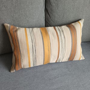Peut inclure: Un coussin rayé avec des rayures brunes, beiges et dorées. Le coussin est sur un canapé gris.