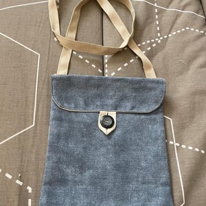 Op de afbeelding: Een crossbodytas in denimstijl met een beige band. De tas heeft een klepsluiting die met een knoop is vastgezet. De tas is blauw van kleur.