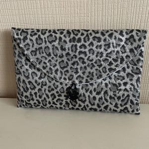 Op de afbeelding: Een rechthoekige clutch met een zilver-zwart luipaardprintpatroon. De klepsluiting is vastgezet met een zwarte knoop. De clutch ligt op een wit oppervlak.