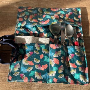 May include: A teal fabric roll-up utensil holder with a colourful ginkgo leaf pattern. The holder contains a spoon, fork, knife, and a small stack of napkins. The knife has a red handle. A dark blue strap is attached.