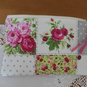 Pode incluir: Uma bolsa branca com zíper com um design de patchwork com rosas cor-de-rosa e folhas verdes. A bolsa tem uma fita rosa presa ao zíper.